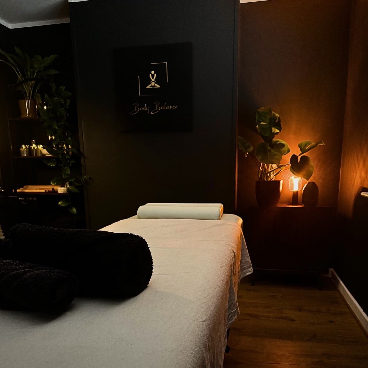 A cozy, dimly lit massage room featuring a neatly prepared massage table with a light-colored sheet and rolled black towels. In the background, soft warm lighting from a lamp and candles creates a relaxing atmosphere, complemented by green plants and minimalist decor. A wall sign reading “Body Balance” adds a professional spa-like touch.